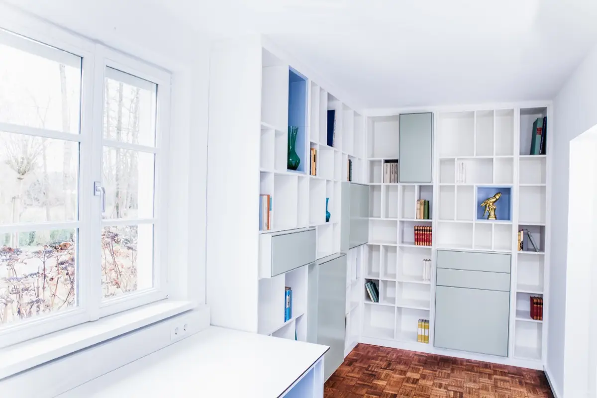 Arbeitszimmer mit umlaufender Regalwand aus dem Regalsystem H, davor ein Fenster mit Blick ins Grüne