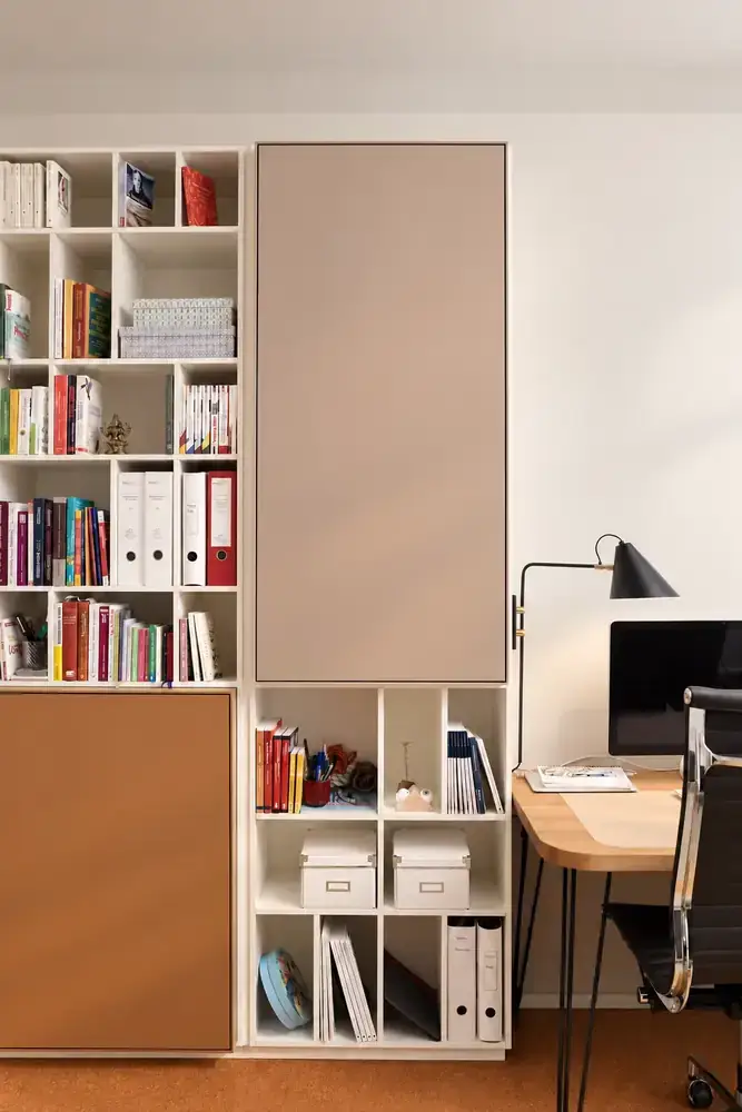 Raumhohe Regalwand aus dem Regalsystem H mit großen Klappen in Taupe und Cognac, daneben ein Schreibtisch mit Lampe