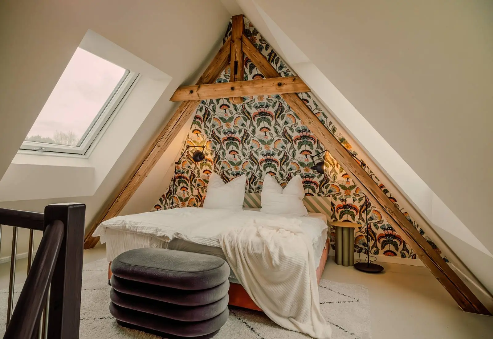 Schlafzimmer unter Dachbalken mit gemusterter Tapete und Samthocker in einer Ferienwohnung der Villa Haag