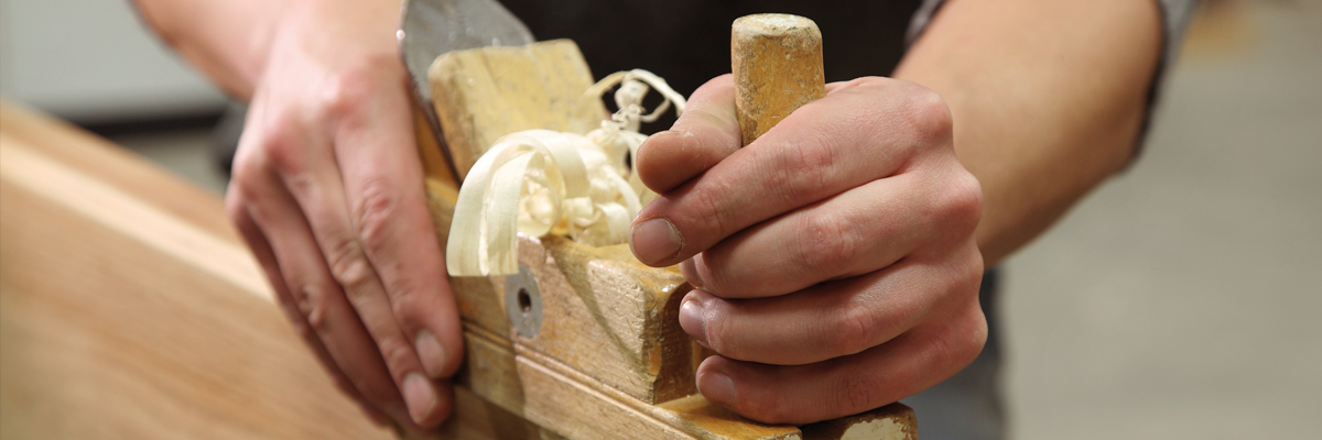 Hände führen einen Hobel über ein Werkstück aus Holz in der Schreinerwerkstätte Weber